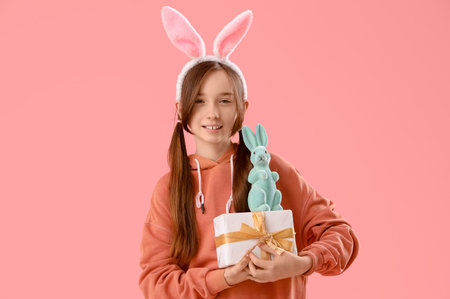 Cute little girl with Easter bunny ears, gift and toy rabbit on pink background
