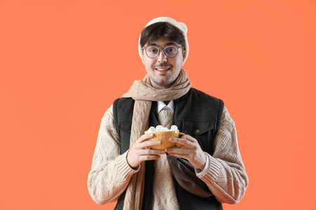 Young man in winter clothes with cup of hot cocoa on orange background