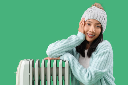 Young Asian woman in winter clothes with radiator on green backgroundの写真素材