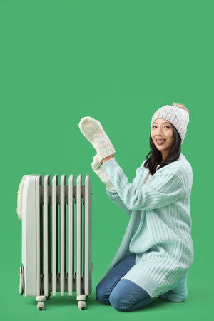 Young Asian woman in winter clothes with radiator on green backgroundの写真素材
