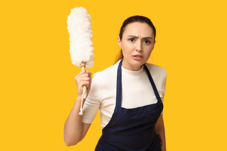 Portrait of angry young housewife in apron with pp duster on yellow backgroundの写真素材