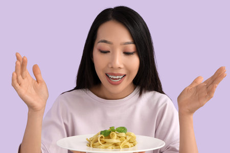 Young Asian woman with tasty pasta on lilac background, closeup