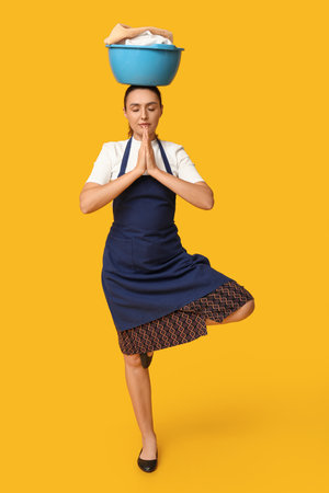 Young housewife in apron meditating with basket of dirty laundry on her head against yellow backgroundの写真素材