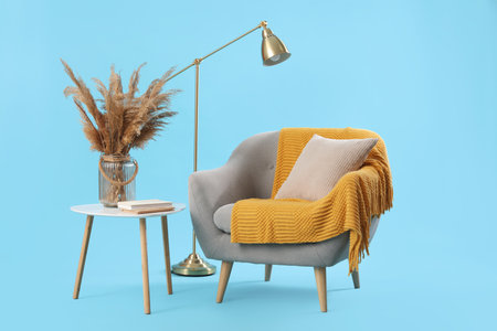 Cozy gray armchair with cushion, warm blanket and pampas grass on coffee table against blue backgroundの写真素材