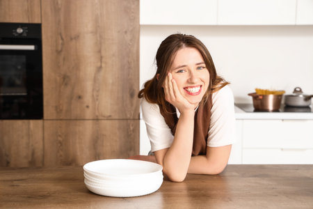 Pretty young woman leaning on table with clean dishes in modern kitchenの写真素材