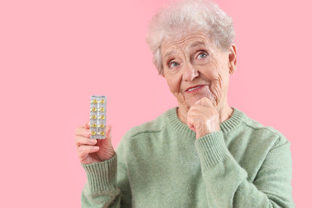 Thoughtful senior woman with pills on pink background, closeup