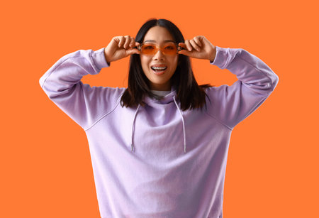 Young Asian woman in lilac hoodie with sunglasses on orange backgroundの写真素材