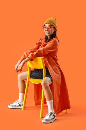 Stylish young Asian woman sitting in chair on orange backgroundの写真素材