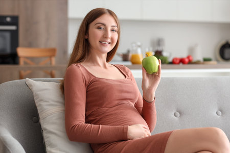 Young pregnant woman with apple sitting on sofa at home
