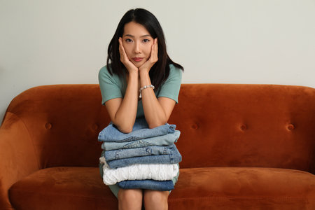 Tired young woman with stack of clean clothes sitting on brown sofaの写真素材