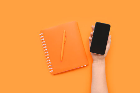 Female hand holding smartphone with blank screen and notebook on orange backgroundの写真素材