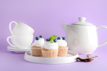 Plate of tasty cupcakes with blueberries, teapot and cups on lilac backgroundの写真素材