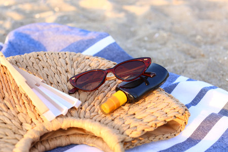 Stylish sunglasses with straw bag and sunscreen on sandy background