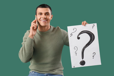 Young man with paper question marks talking by mobile phone on green backgroundの写真素材