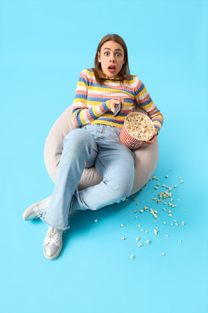 Shocked young woman with popcorn watching movie on blue backgroundの写真素材