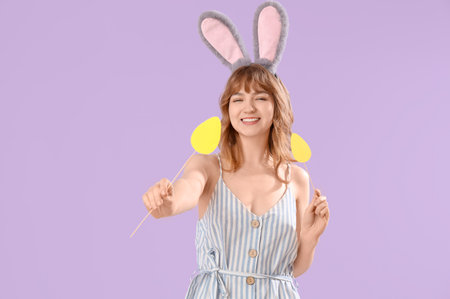 Young girl with Easter bunny ears and paper eggs on lilac background