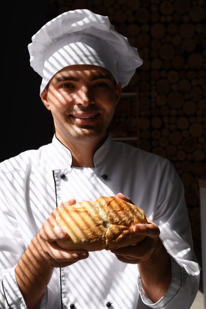 Young chef with loaf of fresh bread in bakery near windowの写真素材