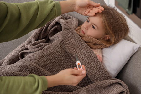 Mother with thermometer touching forehead of her sick little girl at home