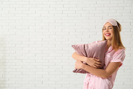 Happy young woman in sleep mask with pink soft pillow on white brick backgroundの写真素材