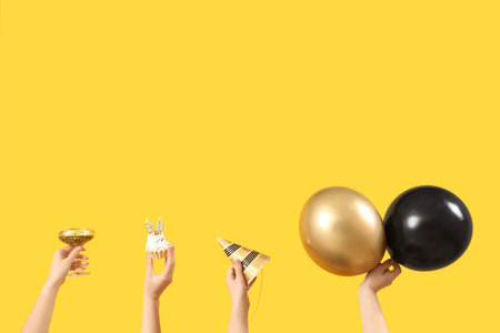 Female hands with balloons, glass, cupcake and party cone on yellow backgroundの写真素材