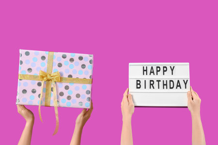 Female hands holding gift box and board with text HAPPY BIRTHDAY on purple backgroundの写真素材