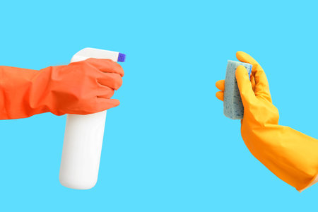 Female hands in rubber gloves holding bottle of detergent and sponge on blue background
