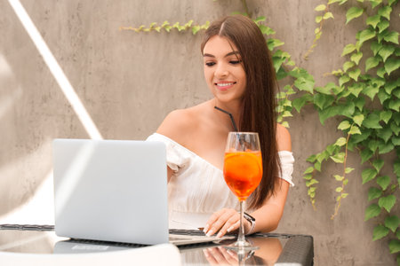Beautiful young Asian woman with glass of tasty  spritz and laptop sitting in cafe, outdoorsの写真素材