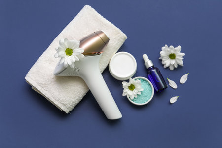 Modern photoepilator with beautiful flowers, bath towel and cosmetic products on dark blue backgroundの写真素材