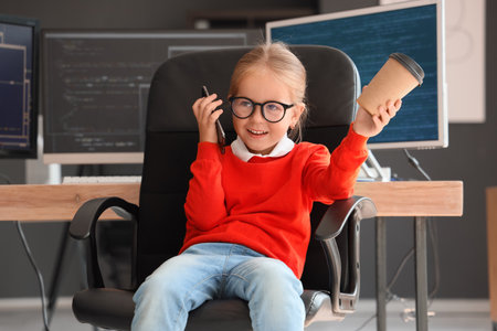 Cute little programmer girl with cup of coffee talking by phone in officeの写真素材