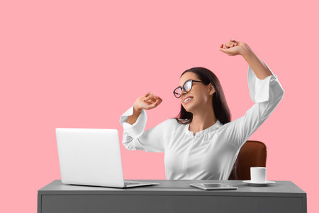 Tired young female programmer stretching at table with laptop on pink backgroundの写真素材