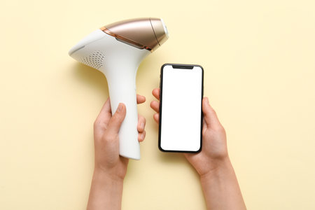 Female hands with mobile phone and modern photoepilator on beige backgroundの写真素材