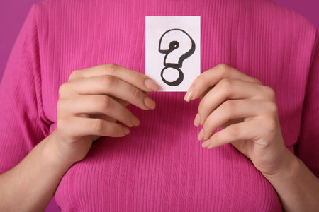 Thoughtful young woman holding paper sheet with question mark on purple background