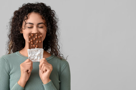 Beautiful young African-American woman with sweet chocolate bar on gray background