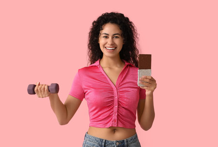 Happy young African-American woman with sweet chocolate bar and dumbbell on pink background
