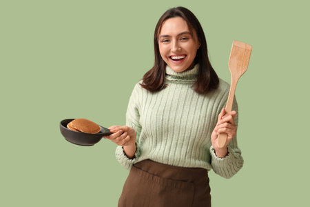 Happy young woman holding frying pan with tasty pancakes on green backgroundの写真素材