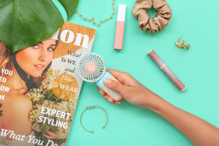 Female hand with portable electric fan, fashion magazine and jewelery on turquoise background