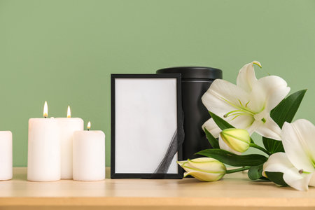 Blank funeral frame, burning candles and beautiful lily flowers on wooden table near color wallの写真素材