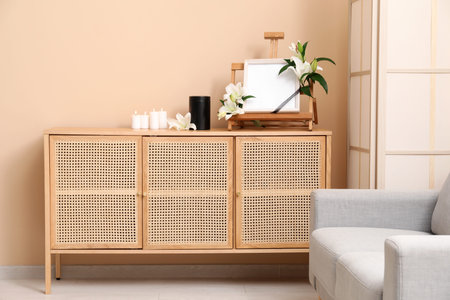 Blank funeral frame with lily flowers, mortuary urn and burning candles on wooden cabinet against color wallの写真素材