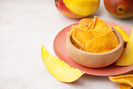 Wooden bowl with slices of dried mango and fresh fruit on white tableの写真素材