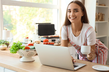 Beautiful young woman using laptop while cooking delicious sausages on modern electric grill in kitchenの写真素材
