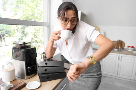 Busy young businesswoman drinking coffee while talking by phone in kitchenの写真素材
