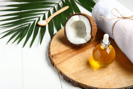 Decorative stand with bottle of coconut cosmetic oil and bath towel on white wooden background
