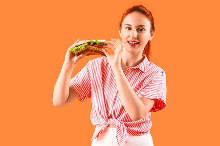 Young redhead woman with tasty sandwich on orange backgroundの写真素材