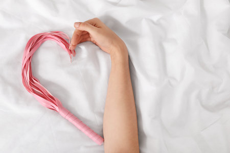 Woman with whip from sex shop making heart gesture on white blanket