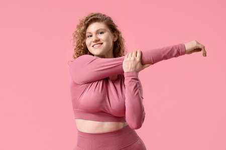 Sporty young woman stretching arms on pink background