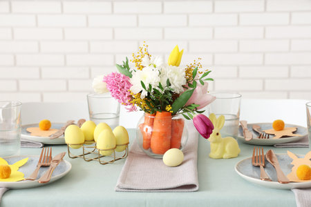 Beautiful table setting for Easter decorated with bouquet of carrots and flowers near white brick...
