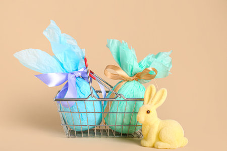 Small shopping basket with wrapped Easter eggs and bunny figurine on beige background