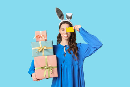 Happy young woman in Easter bunny ears headband with gift boxes and credit card on blue background