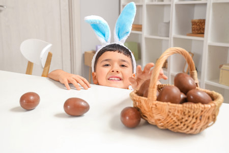 Cute little boy in bunny ears headband with basket of chocolate eggs for Easter at home