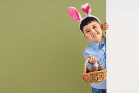 Cute little boy in bunny ears headband holding basket with chocolate eggs for Easter on green...